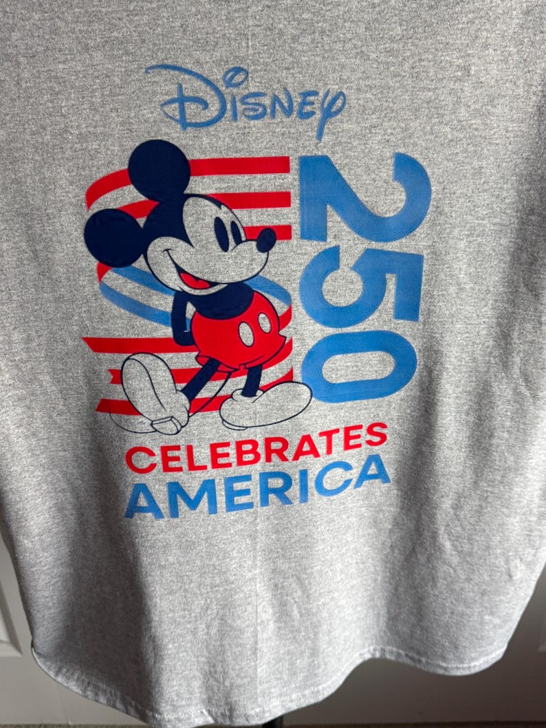 May include: A heather grey t-shirt featuring a graphic of Mickey Mouse in red shorts. The design includes the blue text Disney, the number 250 in large blue font, and the red and blue text CELEBRATES AMERICA over red and blue wavy stripes.