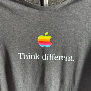 May include: Dark gray t-shirt featuring a colorful apple logo and the text "Think different." The apple logo is a rainbow design. The shirt has a V-neck and is made of a soft material.