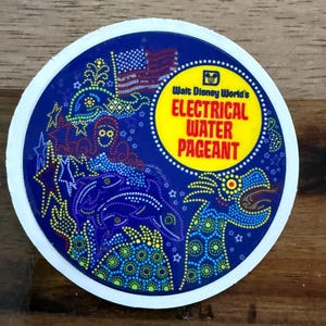 May include: A round sticker with a blue background and a white border. The sticker features the text "Walt Disney World's ELECTRICAL WATER PAGEANT" in yellow and red. The design includes stylized aquatic creatures and an American flag.