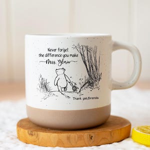 May include: A white ceramic mug with a speckled finish and a tan base. The front features a black line drawing of Winnie the Pooh and Piglet walking, with the text: Never forget the difference you make Mrs. Gloria Thank you, Brenda.