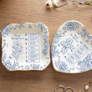 Può includere: Due piatti decorativi in ceramica per anelli, uno quadrato e uno a forma di cuore, con disegni floreali blu e bordi dorati. I piatti riportano la scritta "God Says I Am". Sono visibili anche una collana d'oro, anelli e penne.