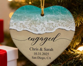 Könnte beinhalten: Herzförmiger Ornament mit Strandmotiv. Der obere Teil zeigt türkisfarbenes Wasser und weiße Wellen, die in einen Sandstrand übergehen. Das Wort "engaged" ist in Schrift geschrieben, zusammen mit Namen und einem Datum. Ein goldener Faden ist zum Aufhängen befestigt.