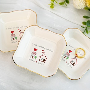 以下が含まれることがあります： 金色の縁取りが付いた、3つのオフホワイトのセラミック製の小物入れ。 各皿には、ハートと「I wish you lived next door」のテキストが入った家のグラフィックが描かれています。「William」と「Sophia」の名前も印刷されています。