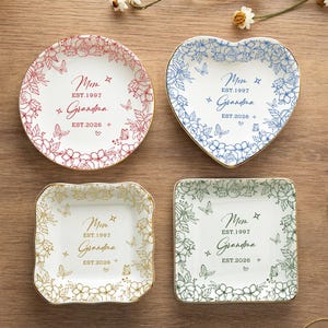 Könnte beinhalten: Vier dekorative Schmuckschalen in verschiedenen Formen: rund, herzförmig und quadratisch. Jede Schale zeigt Blumen- und Schmetterlingsmuster mit dem Text "Mom Grandma EST. 1997 EST. 2026". Die Schalen haben goldfarbene Ränder und sind in Rot-, Blau-, Gold- und Grüntönen gehalten.