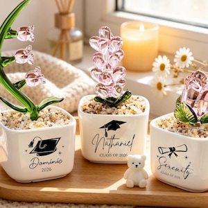 May include: Three white square ceramic planters filled with small pebbles, each featuring a glass flower and black text. The first shows a lily of the valley, a graduation cap, and the name Dominic 2026. The second has a pink flower, a cap, and Nathaniel Class of 2026. The third has a rose, a diploma, and Serenity Class of 2026.