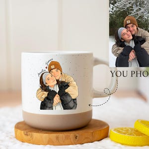 Puede incluir: Una taza de cerámica blanca con un diseño moteado y una base beige. La taza presenta una ilustración personalizada de una pareja abrazándose, con los nombres "Amelia" y "James" escritos encima. A la derecha se muestra una foto de la pareja.