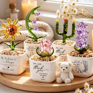 May include: A collection of glass flower sculptures in small white pots, including a sunflower, lily, rose, cactus, and hyacinth. Each pot has a message and sits on a wooden tray with a small teddy bear.
