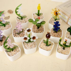 以下が含まれることがあります： 小さな白い正方形の鉢に入った、ミニチュアガラスの花の彫刻のコレクション。花は紫、黄色、青、緑など様々な色があります。鉢には小さな小石が詰められており、繊細で芸術的な雰囲気を醸し出しています。