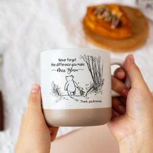 May include: A white ceramic mug with a speckled finish and a tan base. The front features a black line drawing of Winnie the Pooh and Piglet walking, with the text: Never forget the difference you make Mrs. Gloria. Thank you, Brenda.
