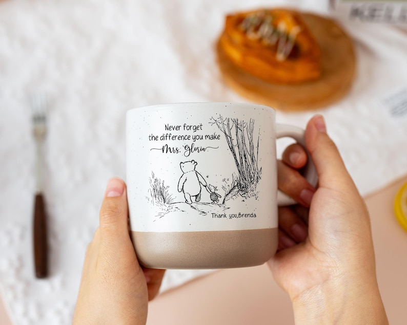 May include: A white ceramic mug with a speckled finish and a tan base. The front features a black line drawing of Winnie the Pooh and Piglet walking, with the text: Never forget the difference you make Mrs. Gloria. Thank you, Brenda.