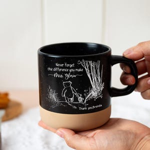 May include: A black ceramic mug with a speckled finish and a tan base. The mug features a white line-art illustration of two characters walking in a forest, with the text "Never forget the difference you make Mrs. Gloria" and "Thank you, Brenda."
