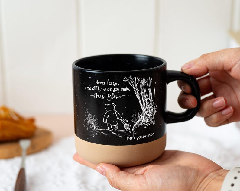 May include: A black ceramic mug with a speckled finish and a tan base. The mug features a white line-art illustration of two characters walking in a forest, with the text "Never forget the difference you make Mrs. Gloria" and "Thank you, Brenda."