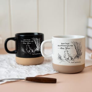 May include: Two ceramic mugs, one black and one white, featuring a speckled finish and a tan base. Each mug displays a line art illustration of a bear and a small animal walking in a forest, with the text "Never forget the difference you make Mrs. Gloria" and "Thank you, Brenda."