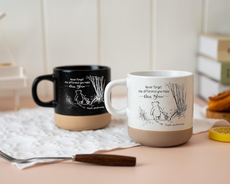 May include: Two ceramic mugs, one black and one white, featuring a speckled finish and a tan base. Each mug displays a line art illustration of a bear and a small animal walking in a forest, with the text "Never forget the difference you make Mrs. Gloria" and "Thank you, Brenda."