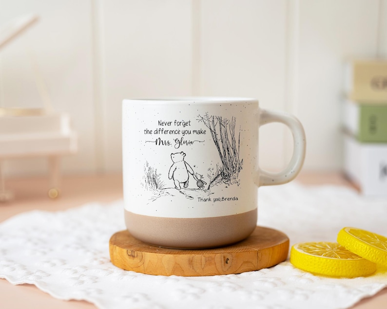 May include: A white ceramic mug with a speckled finish and a tan base. The front features a black line drawing of Winnie the Pooh and Piglet walking, with the text: Never forget the difference you make Mrs. Gloria Thank you, Brenda.