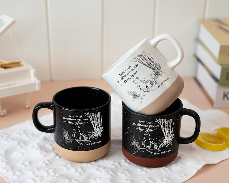 May include: Three ceramic mugs featuring a white line art illustration of a bear walking in a forest. Each mug displays the text 'Never forget the difference you make', 'Mrs. Glow', and 'Thank you Brenda'. Two mugs are black, one is white.