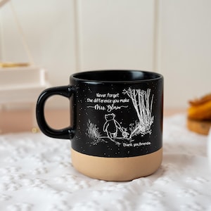 May include: A black ceramic mug with a speckled finish and a tan base. The front features a white line drawing of two characters walking near trees, with the text "Never forget the difference you make Mrs. Gloria" and "Thank you, Brenda."