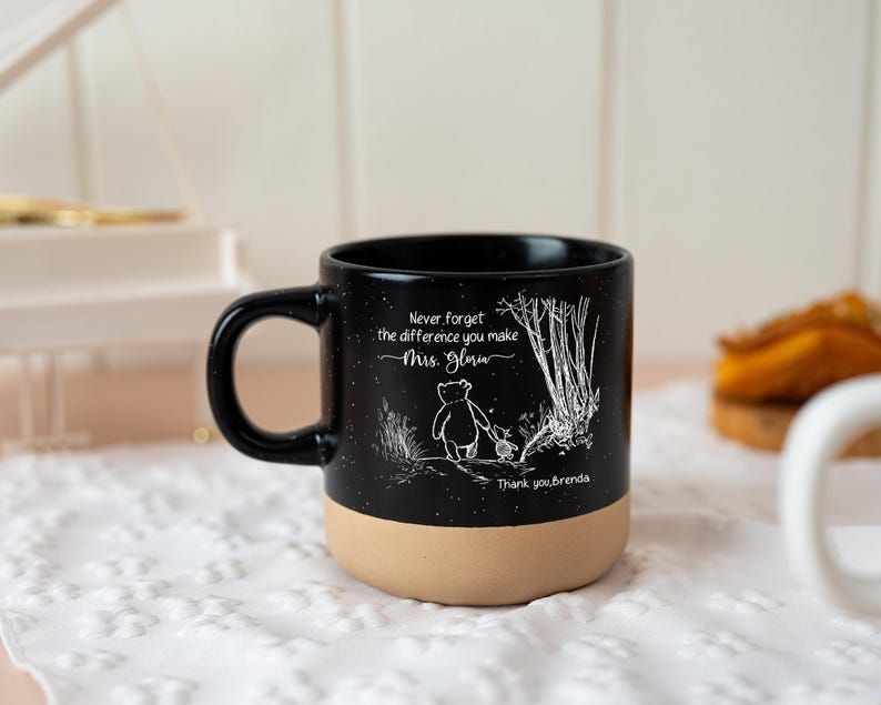 May include: A black ceramic mug with a speckled finish and a tan base. The front features a white line drawing of two characters walking near trees, with the text "Never forget the difference you make Mrs. Gloria" and "Thank you, Brenda."