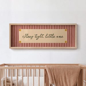 Könnte beinhalten: Ein rechteckiges Wandschild in einem hellen Holzrahmen mit einem cremefarbenen Mittelteil, das den Text "Sleep tight, little one" in dunkler Schrift zeigt, umgeben von einem rosa-braunen vertikal gestreiften Rand mit kleinen Stern- und Mondsymbolen.