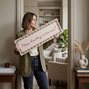 Pu&ograve; includere: Un cartello rettangolare con la scritta "How lucky are we?" in corsivo rosa. Il cartello ha uno sfondo chiaro con un bordo a scacchi rosa e verde e una cornice in legno naturale. Il cartello &egrave; tenuto davanti a un interno dai toni neutri.