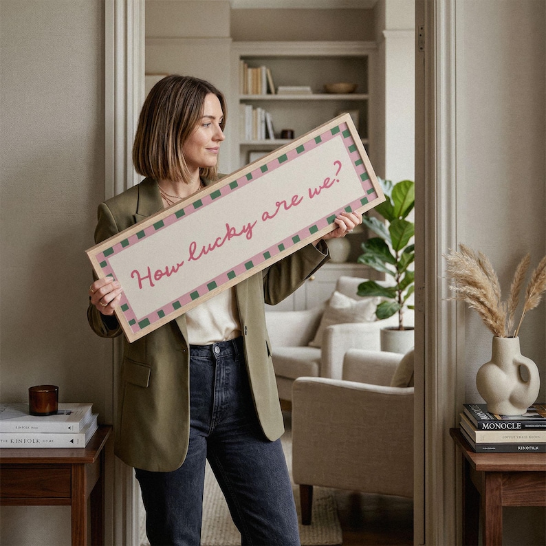 Pu&ograve; includere: Un cartello rettangolare con la scritta "How lucky are we?" in corsivo rosa. Il cartello ha uno sfondo chiaro con un bordo a scacchi rosa e verde e una cornice in legno naturale. Il cartello &egrave; tenuto davanti a un interno dai toni neutri.