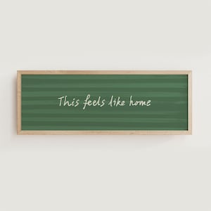 May include: A rectangular wooden framed wall art piece. The artwork features a green background with horizontal lines and the handwritten text "This feels like home" in white cursive lettering.