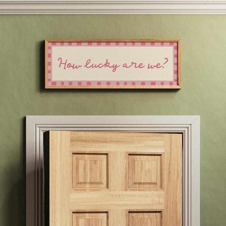 Pu&ograve; includere: Un cartello rettangolare incorniciato con la scritta "How lucky are we?" in corsivo rosa. Il cartello ha un bordo a scacchi rosa e bianco e una cornice in legno chiaro. &Egrave; montato su una parete verde sopra una porta di legno.