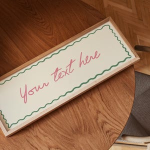 May include: A rectangular wooden framed sign with the text "Your text here" in pink script. The sign has a cream-coloured background with a green wavy border. The sign is resting on a wooden table.