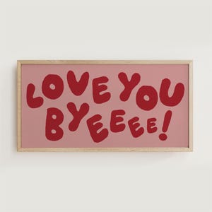 Może przedstawiać: Oprawiony wydruk artystyczny z napisem "LOVE YOU BYEEEE!" w czerwonych, zaokrąglonych literach na różowym tle. Rama jest w jasnym kolorze drewna, a grafika jest wyśrodkowana.