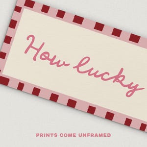 Pu&ograve; includere: Stampa rettangolare con sfondo color crema e bordo rosa con quadrati rosso scuro. Le parole "How lucky" sono scritte in rosa in corsivo. Il testo "PRINTS COME UNFRAMED" &egrave; in basso.