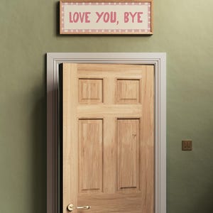 May include: A wooden door with a gold door handle and a sign above it that reads "LOVE YOU, BYE" in pink letters on a white background with a pink and white checkered border.