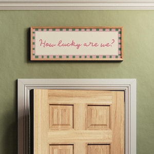 Pu&ograve; includere: Un cartello rettangolare incorniciato con la scritta "How lucky are we?" in corsivo. Il cartello ha un bordo a scacchi rosa e verde ed &egrave; montato su un muro verde sopra una porta di legno.