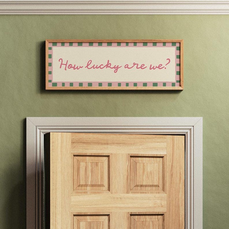 Pu&ograve; includere: Un cartello rettangolare incorniciato con la scritta "How lucky are we?" in corsivo. Il cartello ha un bordo a scacchi rosa e verde ed &egrave; montato su un muro verde sopra una porta di legno.