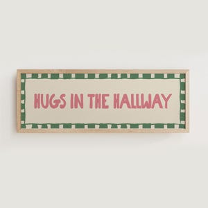 Könnte beinhalten: Rechteckiges Wandbild mit dem Text "HUGS IN THE HALLWAY" in Rosa. Der Text ist auf einem cremefarbenen Hintergrund zentriert, von einem grün-weißen Rand umgeben und in einem Holzrahmen eingefasst.