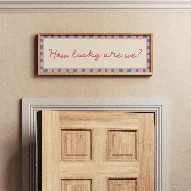 Pu&ograve; includere: Un'opera d'arte rettangolare incorniciata con la scritta "How lucky are we?" in rosa. L'opera d'arte ha uno sfondo beige chiaro con un bordo a scacchi rosa e blu. La cornice &egrave; di colore marrone chiaro.
