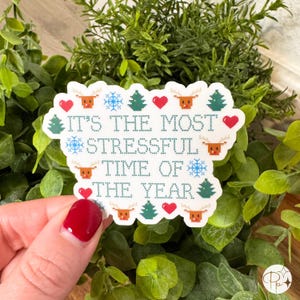 A glossy vinyl sticker designed like a holiday cross-stitch pattern with the phrase “It’s the Most Stressful Time of the Year.” Surrounded by pixel-style reindeer, snowflakes, Christmas trees, and red hearts, the design humorously captures the chaos of the holidays. Perfect for adding festive humor to planners, notebooks, or gift wrapping. Measures approximately 2.8 inches wide by 2.1 inches tall, available in glossy finish.