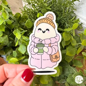May include: A sticker featuring a cute cartoon character in a pink winter coat and a knit hat with a pom-pom. The character is holding a green coffee cup and a tan purse. The sticker has a white border and is set against a background of green foliage.
