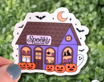May include: A die-cut sticker featuring a purple bookstore with a brown roof, orange pumpkins, and ghosts. The sign reads Spooky Bookstore. The illustration includes bats, a crescent moon, and bookshelves visible through the windows.