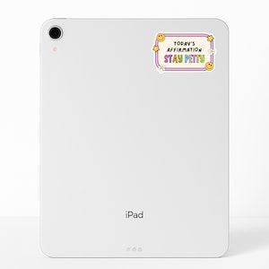 May include: A white iPad with a sticker that reads "Today's Affirmation Stay Petty" in a retro design. The sticker features a pink border, yellow stars, smiley faces, and white flowers. The iPad has a camera and the word "iPad" printed on the bottom.