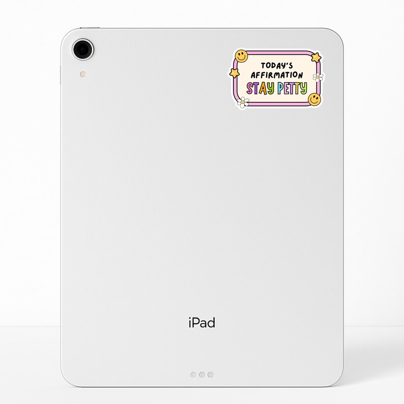 May include: A white iPad with a sticker that reads "Today's Affirmation Stay Petty" in a retro design. The sticker features a pink border, yellow stars, smiley faces, and white flowers. The iPad has a camera and the word "iPad" printed on the bottom.