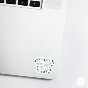 May include: A white sticker on a laptop with the text "IT'S THE MOST STRESSFUL TIME OF THE YEAR" in teal. The sticker is decorated with red hearts, snowflakes, Christmas trees, and reindeer.