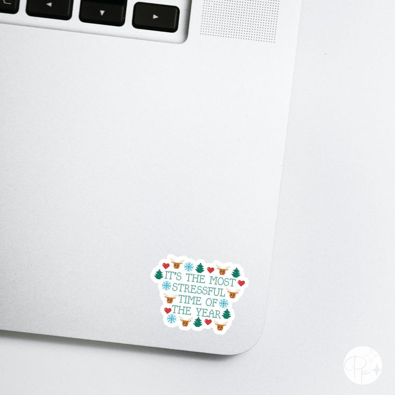 May include: A white sticker on a laptop with the text "IT'S THE MOST STRESSFUL TIME OF THE YEAR" in teal. The sticker is decorated with red hearts, snowflakes, Christmas trees, and reindeer.