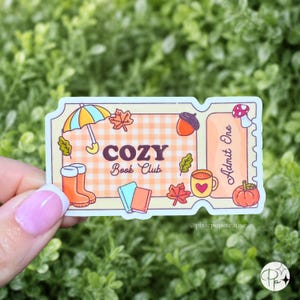 May include: A rectangular sticker shaped like an admission ticket featuring a light orange gingham pattern. The text reads COZY Book Club and Admit One. Illustrations include an umbrella, boots, books, a mug, an acorn, leaves, and a pumpkin.