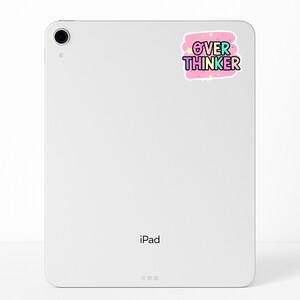 May include: A white tablet with a camera lens in the top left corner and a sticker on the upper right side. The sticker features a pink cloud-shaped background with small stars and the text OVER THINKER in blue and purple bubble letters.