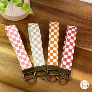 May include: Four key fobs with a checkered pattern in pink, tan, orange, and red. Each fob has a bronze-colored metal clasp and key ring. The fobs are arranged on a wooden surface.