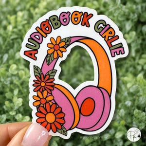 May include: A die-cut sticker featuring a pair of pink and orange headphones adorned with orange flowers and green leaves. The text "AUDIOBOOK GIRLIE" is arched in colorful letters above the headphones. The sticker has a white border.