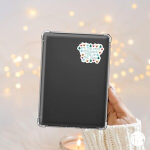 May include: A black tablet with a clear case is held by a person. A sticker on the tablet reads "It's the most stressful time of the year" with festive icons like snowflakes, trees, and hearts in blue, red, and green.
