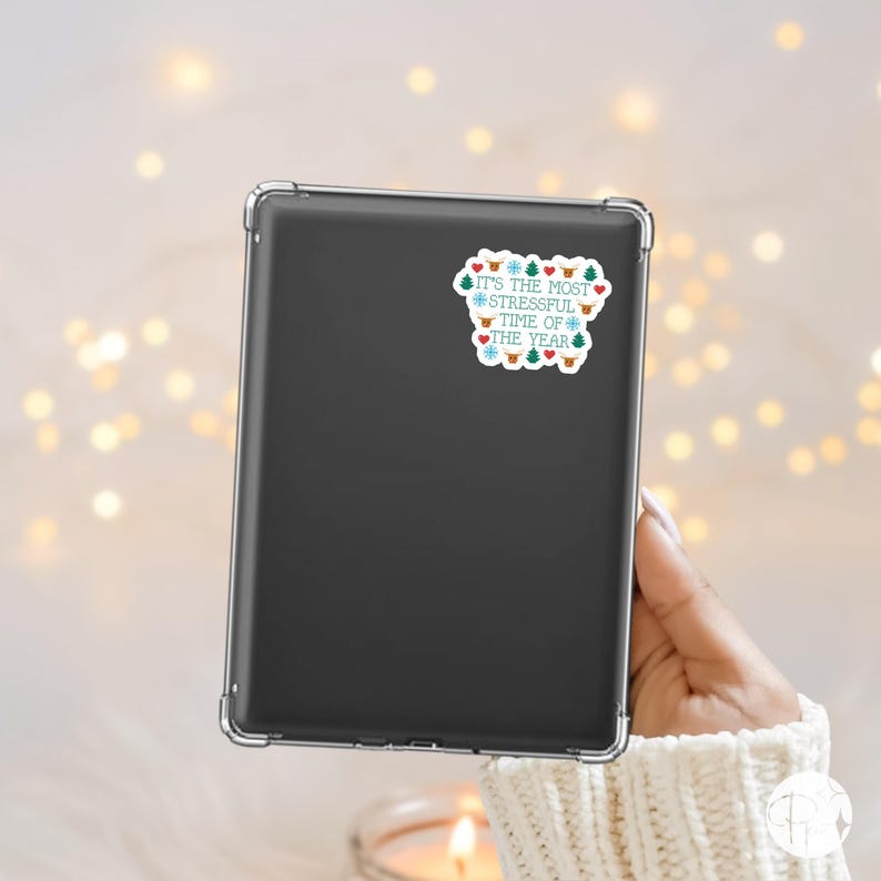 May include: A black tablet with a clear case is held by a person. A sticker on the tablet reads "It's the most stressful time of the year" with festive icons like snowflakes, trees, and hearts in blue, red, and green.