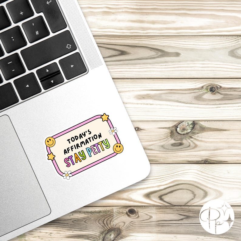 May include: A white sticker on a laptop with the text "TODAY'S AFFIRMATION STAY PETTY" in a pink and yellow frame. The frame has yellow stars, smiley faces, and white flowers. The laptop is on a wooden surface.