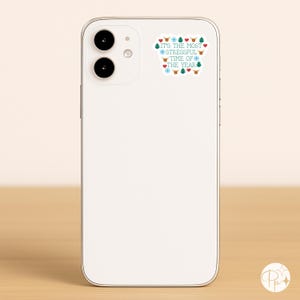 May include: A white smartphone with a sticker that reads "IT'S THE MOST STRESSFUL TIME OF THE YEAR" in blue and green text. The sticker features festive icons like hearts, snowflakes, reindeer, and Christmas trees.